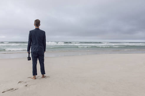Rückenansicht eines Geschäftsmannes, der mit einer VR-Brille am Strand steht, Nordhoek, Westkap, Südafrika - MCF00342