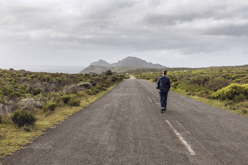 Rückenansicht eines Geschäftsmannes, der auf einer Landstraße spazieren geht, Cape Point, Westkap, Südafrika - MCF00343