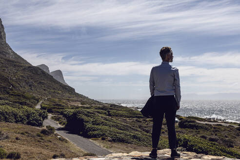 Rückenansicht eines Geschäftsmannes, der auf einem Plateau steht und auf das Meer blickt, Cape Point, Westkap, Südafrika - MCF00348