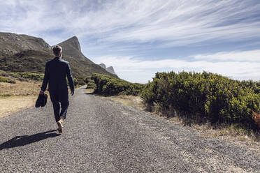 Rückenansicht eines Geschäftsmannes, der barfuß auf einer Landstraße läuft, Cape Point, Westkap, Südafrika - MCF00349