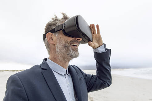 Smiling businessman using VR glasses on the beach, Nordhoek, Western Cape, South Africa - MCF00355