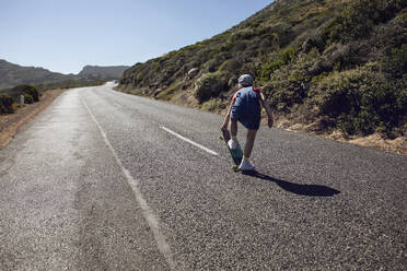 Back view of girl with skateboard standing on country road, Cape Town, Western Cape, South Africa - MCF00374