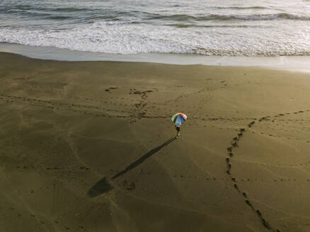 Luftaufnahme einer Person unter Regenbogenfarben Schirm am Strand, Kedungu Strand, Bali, Indonesien - KNTF03732