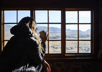 Frau trinkt eine Tasse Kaffee und bewundert die Berge durch das Fenster - VEGF00853