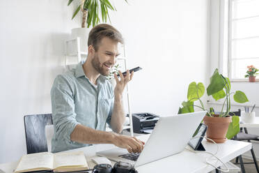 Smiling man using smartphone and laptop at desk in office - VPIF01758