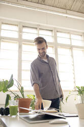 Smiling man looking at book on desk in office - VPIF01790