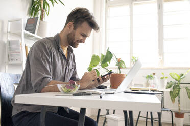 Lächelnder Mann, der während der Mittagspause am Schreibtisch im Büro sein Smartphone benutzt - VPIF01800