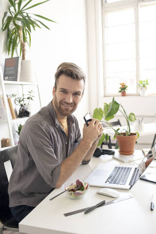 Porträt eines lächelnden Mannes mit Smartphone in der Mittagspause am Schreibtisch im Büro, lizenzfreies Stockfoto
