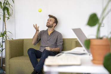 Smiling man sitting on couch in office playing with a tennis ball - VPIF01803