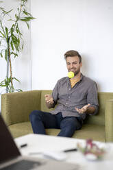 Smiling man sitting on couch in office playing with a tennis ball - VPIF01804