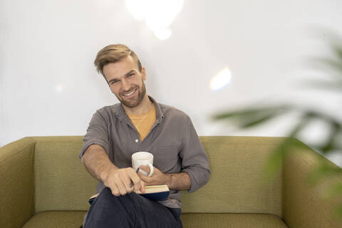 Portrait of smiling man sitting on couch with book and coffee cup stock photo