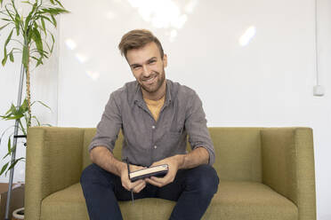 Portrait of smiling man sitting on couch with a book - VPIF01806