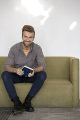 Porträt eines lächelnden Mannes, der mit einem Buch auf der Couch sitzt - VPIF01807