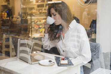 Frau mit Laptop trinkt Kaffee hinter einer Fensterscheibe in einem Cafe - FMOF00760