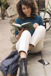 Lächelnde Frau liest ein Buch im Freien in der Stadt - FMOF00784