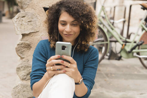 Lächelnde Frau mit Smartphone und Kopfhörern im Freien in der Stadt - FMOF00785