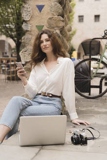 Porträt einer jungen Frau mit Smartphone, Kamera und Laptop im Freien - FMOF00787