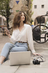 Junge Frau mit Smartphone, Kamera und Laptop im Freien - FMOF00788