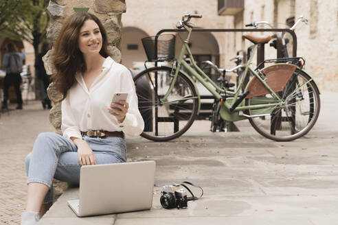 Lächelnde junge Frau mit Smartphone, Kamera und Laptop im Freien - FMOF00789
