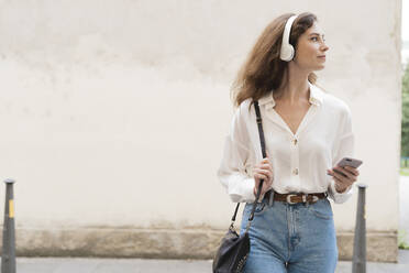 Junge Frau mit Smartphone und Kopfhörern in der Stadt unterwegs - FMOF00801