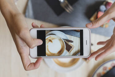 Draufsicht auf eine Frau in einem Café, die mit ihrem Handy einen Cappuccino fotografiert - FMOF00808