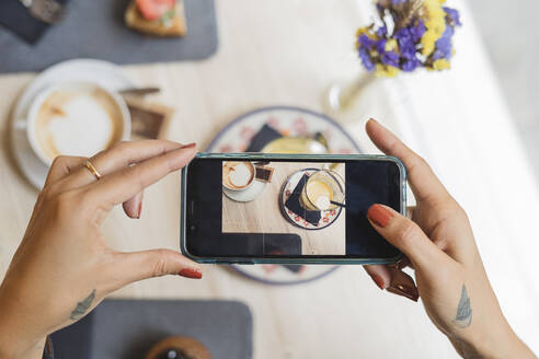 Draufsicht auf eine Frau in einem Café, die mit ihrem Handy einen Kaffee fotografiert - FMOF00809