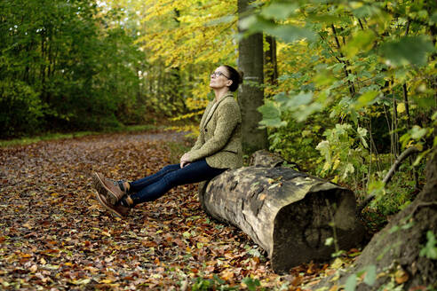 Mature woman sitting on a tree trunk in a autumn forest - FLLF00352