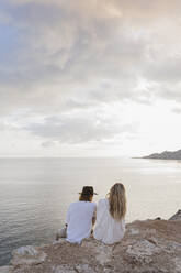 Rückenansicht eines jungen Paares, das auf einem Felsen vor dem Meer sitzt, Ibiza, Balearische Inseln, Spanien - AFVF04272