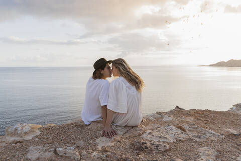 Rückenansicht eines jungen verliebten Paares, das auf einem Felsen vor dem Meer sitzt, Ibiza, Balearische Inseln, Spanien, lizenzfreies Stockfoto