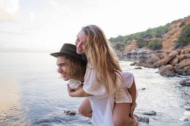 Junger Mann, der seine Freundin vor dem Meer huckepack nimmt, Ibiza, Balearische Inseln, Spanien - AFVF04285