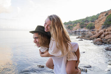 Young man giving his girlfriend a piggyback ride in front of the sea, Ibiza, Balearic Islands, Spain - AFVF04285