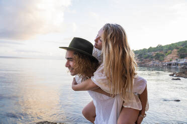 Young man giving his girlfriend a piggyback ride in front of the sea, Ibiza, Balearic Islands, Spain - AFVF04286