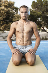 Porträt eines muskulösen Mannes mit nacktem Oberkörper und Kopfhörern, der auf einer Turnmatte kniet - RCPF00149