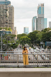 Modische, gelb gekleidete Frau steht auf einer Brücke und schaut auf ihr Handy, Bangkok, Thailand - MAUF03134