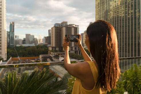 Back view of woman standing on roof terrace taking photo with smartphone, Bangkok, Thailand - MAUF03136