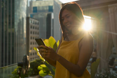 Porträt einer glücklichen Frau auf einer Dachterrasse bei Gegenlicht, die auf ihr Handy schaut - MAUF03148