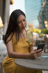 Porträt einer Frau, die mit einem Getränk in einem Café sitzt und auf ihr Mobiltelefon schaut - MAUF03150