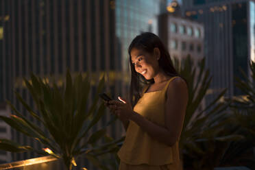 Porträt einer glücklichen Frau, die in der Abenddämmerung auf einer Dachterrasse steht und auf ihr Handy schaut, Bangkok, Thailand - MAUF03155