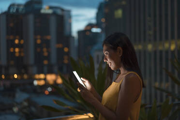 Lächelnde Frau auf einer Dachterrasse in der Abenddämmerung, die auf ihr Mobiltelefon schaut, Bangkok, Thailand - MAUF03158