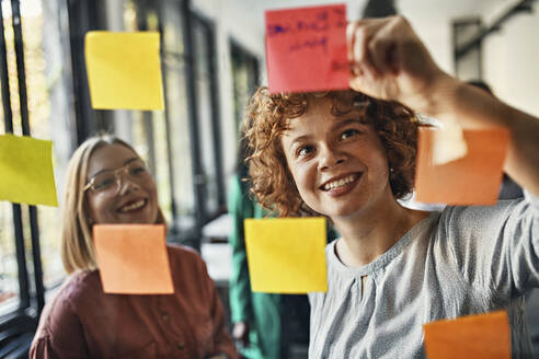 Happy colleagues with sticky notes at glass pane in office - ZEDF02728
