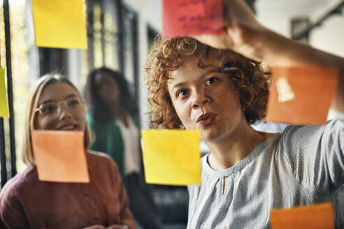 Colleagues writing on sticky notes at glass pane in office - ZEDF02729