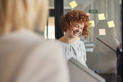 Laughing businesswoman with sticky notes at glass pane in office - ZEDF02733