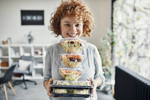 Portrait of smiling businesswoman holding stack of takeaway food in office - ZEDF02792