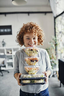 Portrait of smiling businesswoman holding stack of takeaway food in office - ZEDF02793