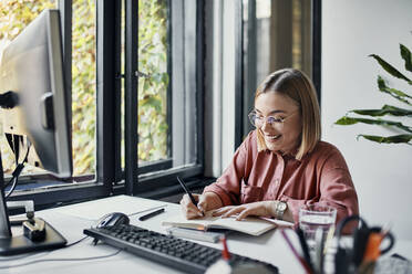 Smiling businesswoman taking notes at desk in office - ZEDF02805