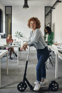Portrait of a smiling businesswoman with kick scooter in office - ZEDF02827