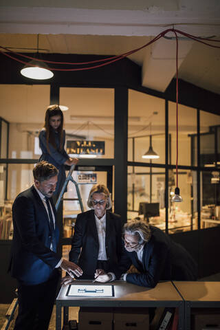 Geschäftsleute, die an einem glänzenden Tablet im Büro diskutieren, mit einem Mädchen auf einer daneben stehenden Leiter, lizenzfreies Stockfoto