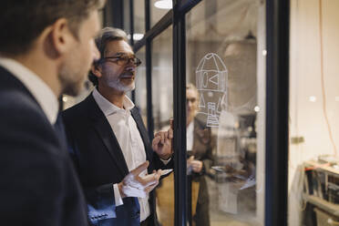 Two businessmen working on drawing on glass pane in office - GUSF02742