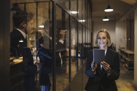 Smiling businesswoman using tablet and two businessmen working on drawing on glass pane in office stock photo