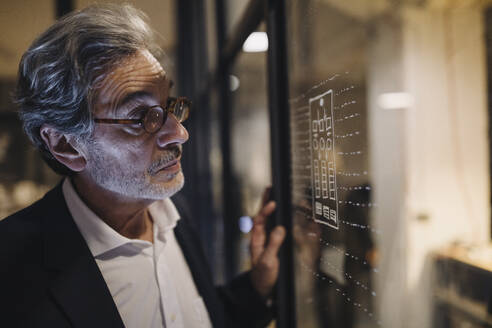 Senior businessman looking at drawing on glass pane in office - GUSF02751